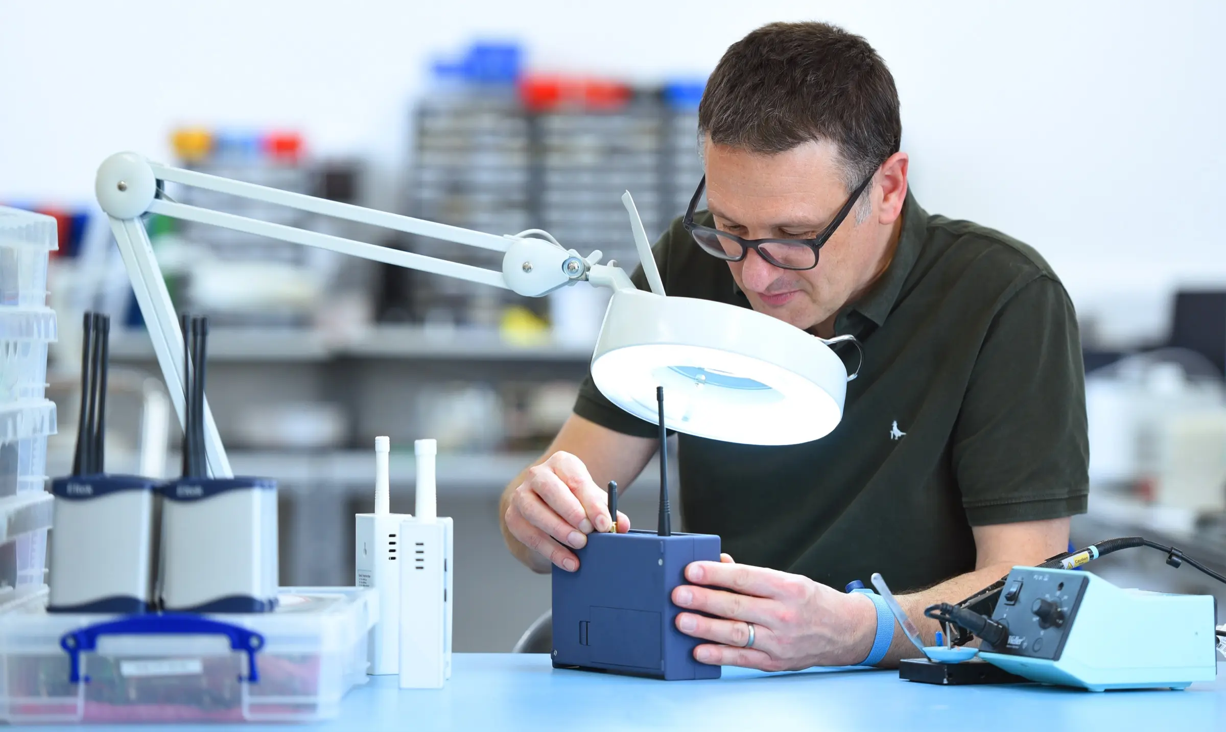 An Eltek engineer inspecting and testing a wireless GenII transmitter under an illuminated magnifier at the UK engineering bench in Royston, the hands-on calibration and build work behind Eltek’s audit-ready monitoring
