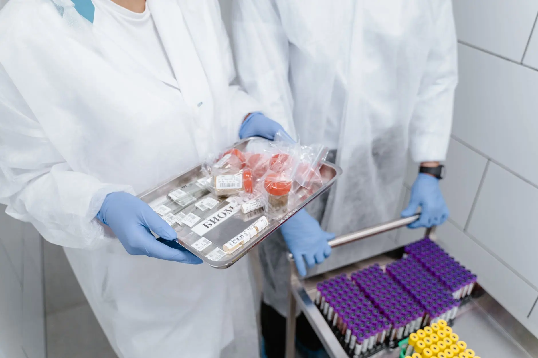 Two gowned laboratory staff handling a tray of labelled patient specimens beside racked blood-collection tubes in a clinical laboratory, the kind of sample handling continuous monitoring safeguards