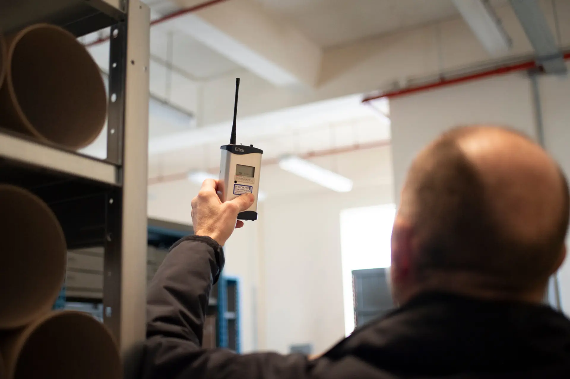 A person raising an Eltek wireless logger to take a reading in an archive store, with overhead building services and ductwork visible, monitoring the environment of a building.