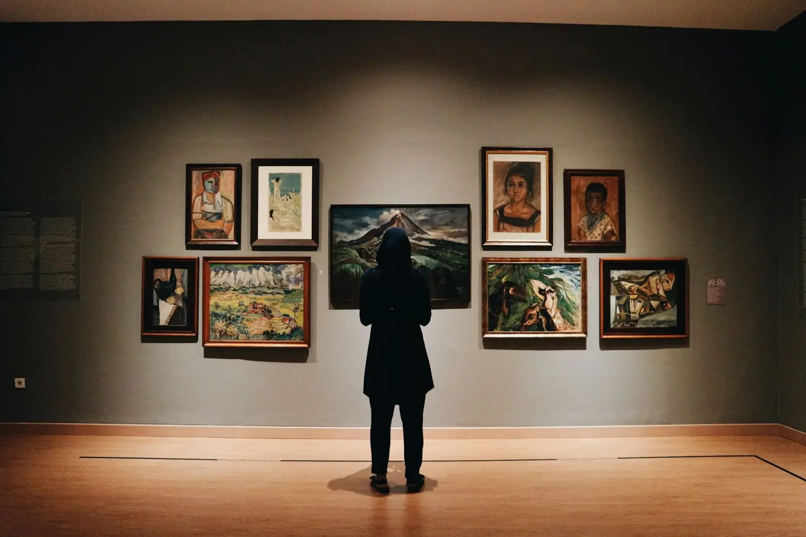 A single visitor alone in a dimly lit exhibition room, facing a close-hung group of framed paintings on a grey gallery wall, the kind of temporary display whose agreed temperature, humidity and light conditions have to be evidenced