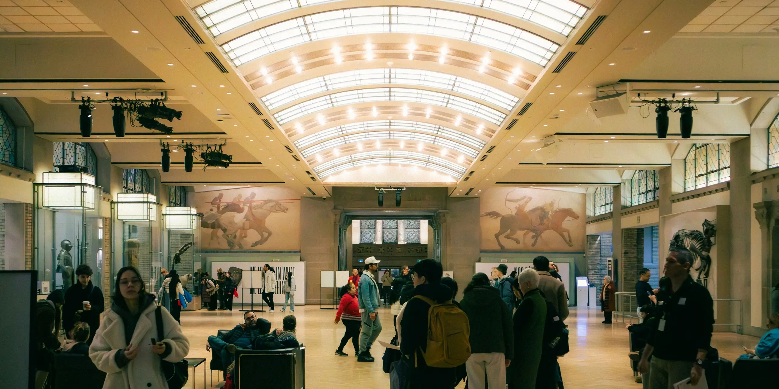 Visitors moving through the top-lit central hall of a public gallery beneath a barrel-vaulted glazed roof and gallery lighting rigs, relief panels on the walls and objects in wall cases, the kind of exhibition space where Eltek evidences the conditions a lender agreed