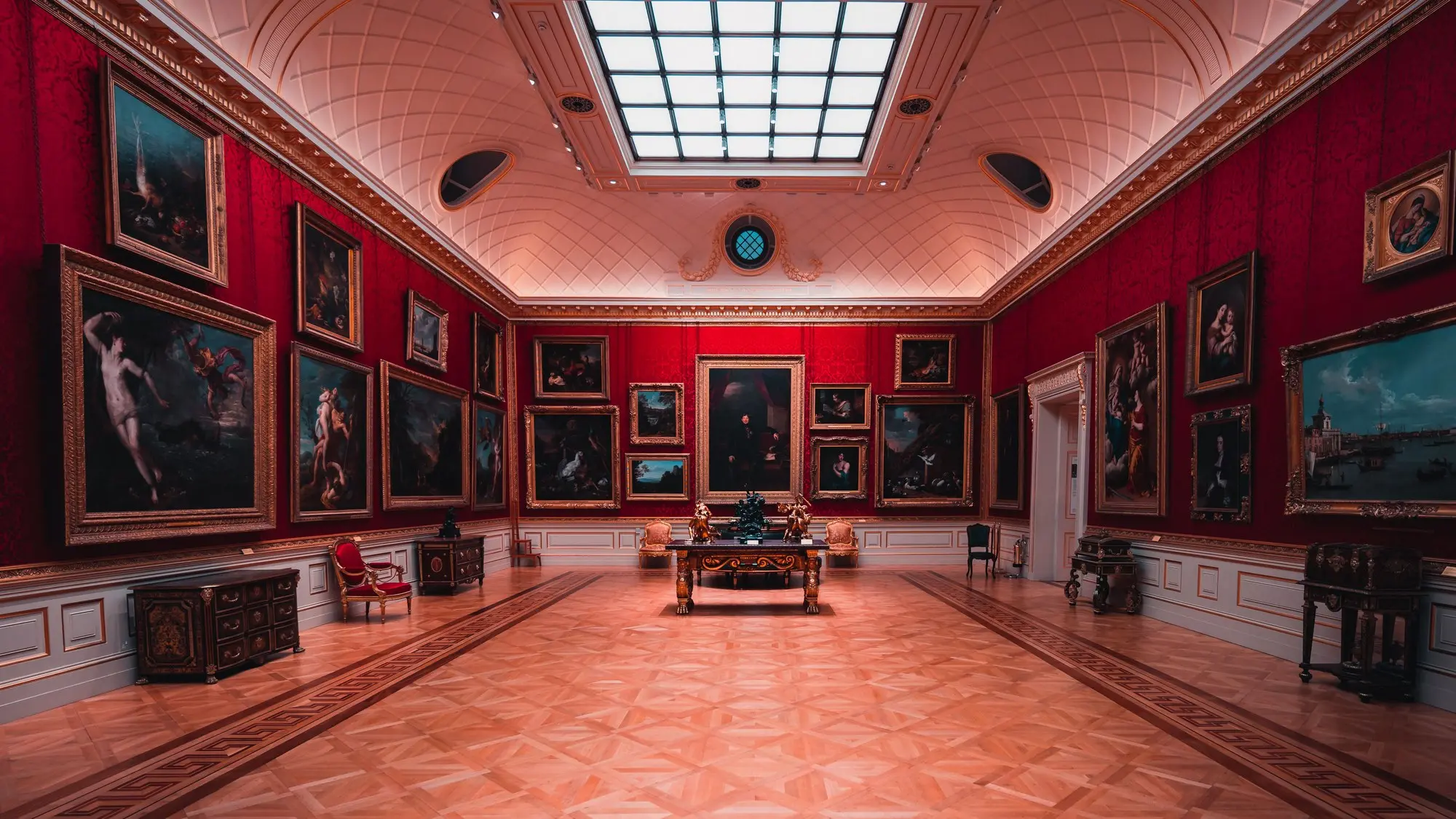 A grand red-walled gallery of framed Old Masters paintings lit from a central roof lantern, the kind of historic interior where a stable environment protects the collection