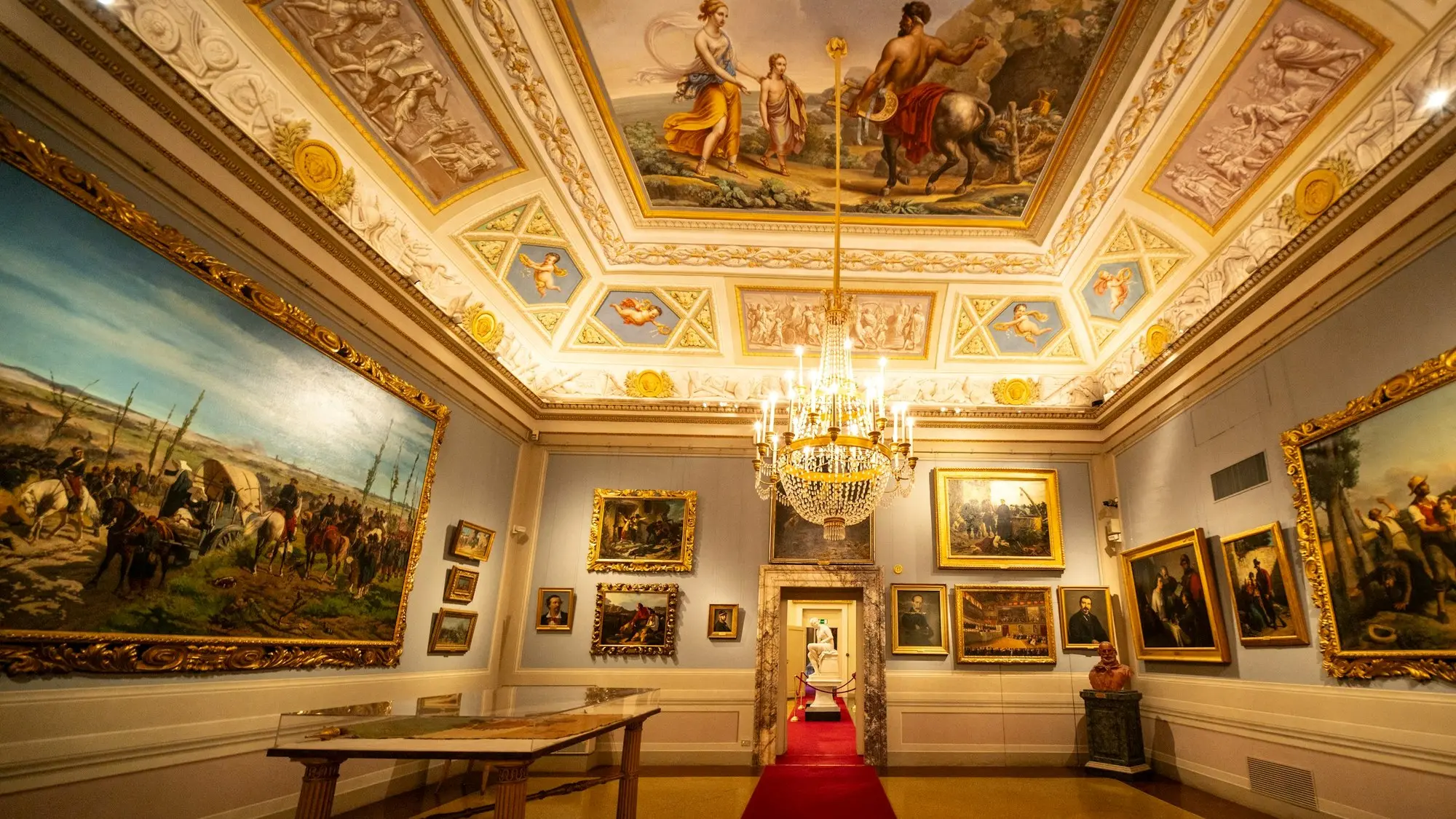 A historic gallery beneath an ornate painted and gilded ceiling, hung with large gilt-framed paintings, the kind of protected decorative fabric that cannot be drilled or chased to run a cable