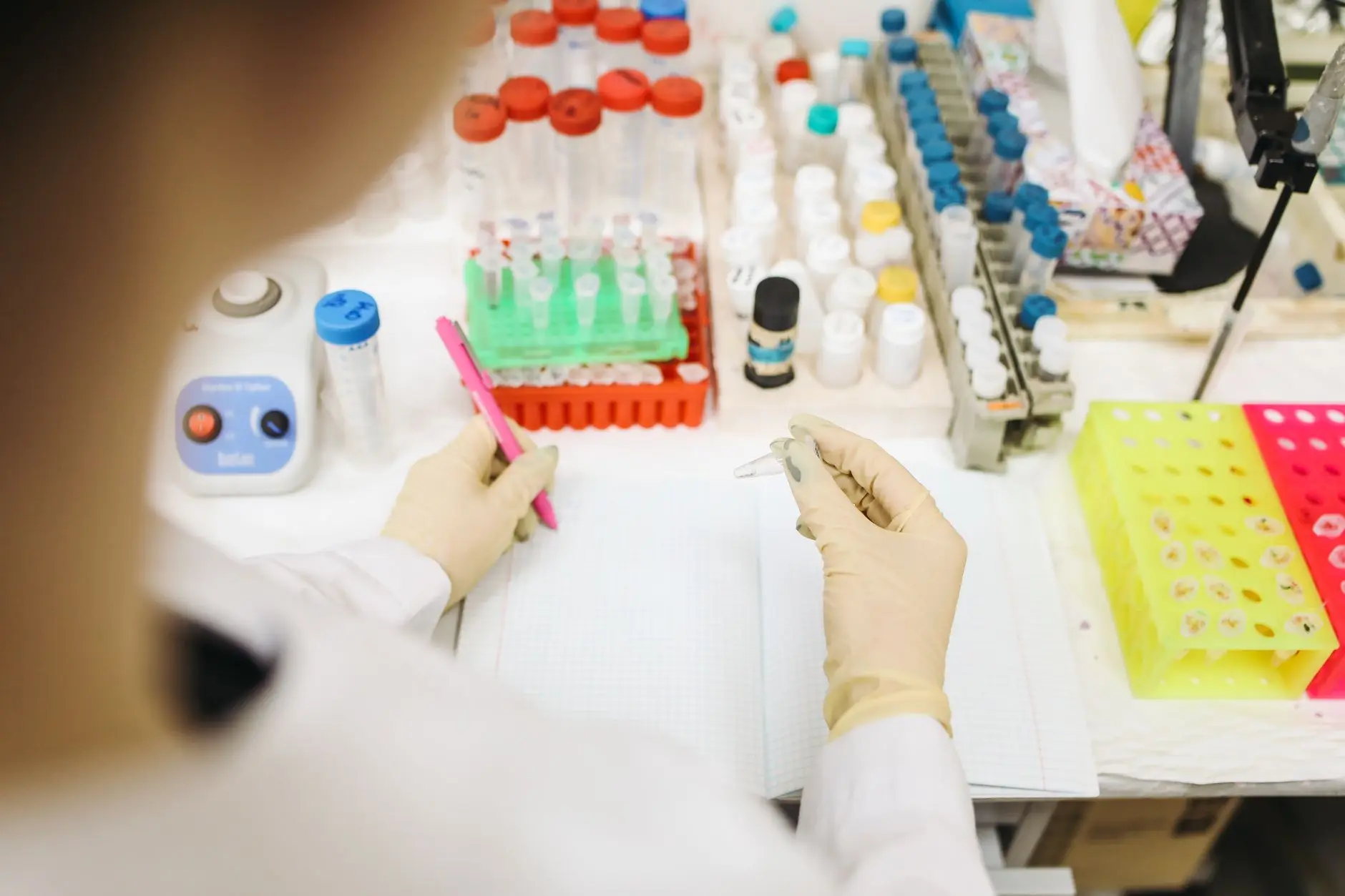 A laboratory technician recording a temperature reading by hand in a paper logbook while handling sample vials at the bench