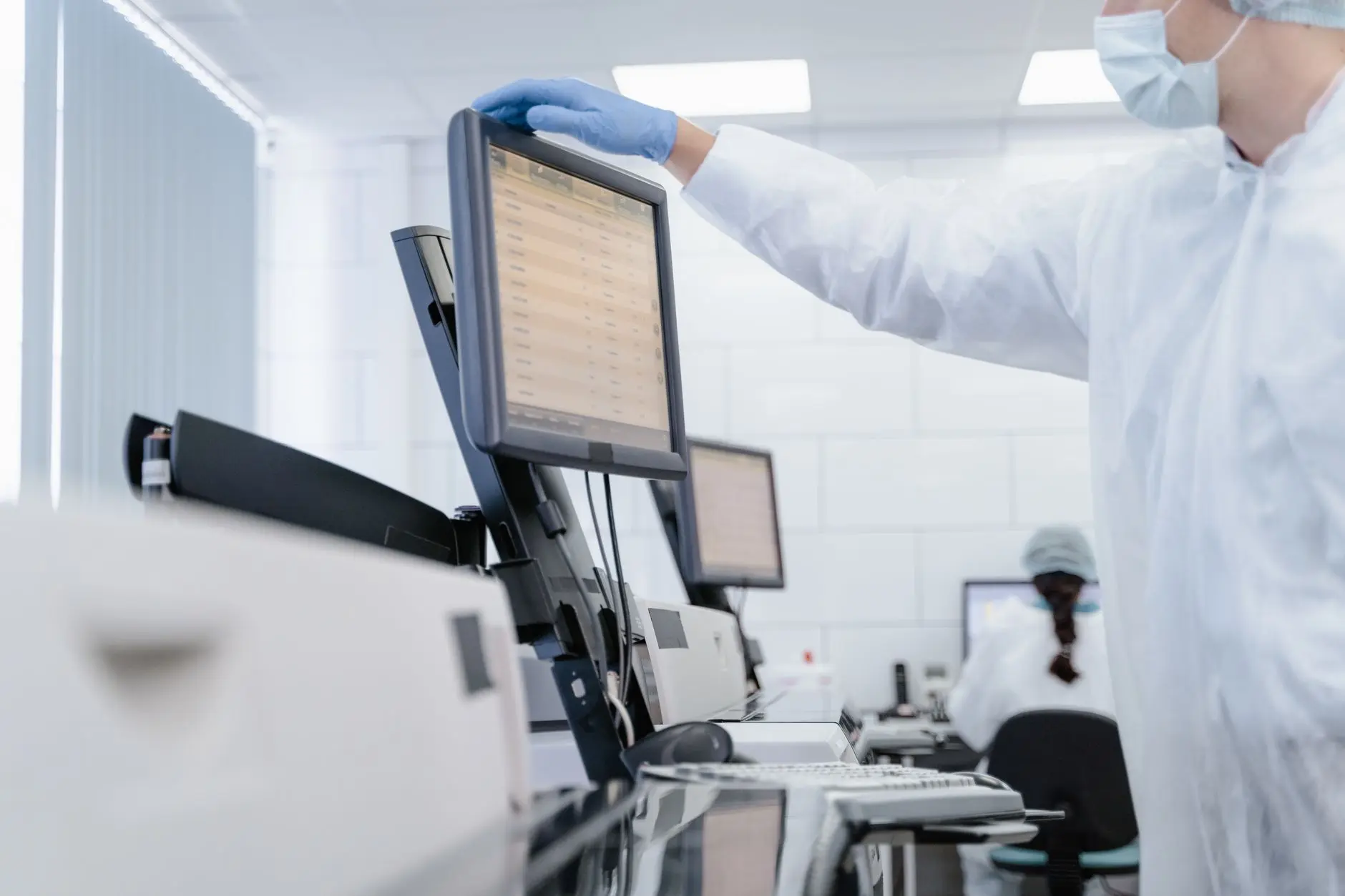 A gowned lab technician reviewing a screen of continuous monitoring readings in a modern life-science laboratory, the kind of verifiable, timestamped data record that supports both compliance and sustainability reporting