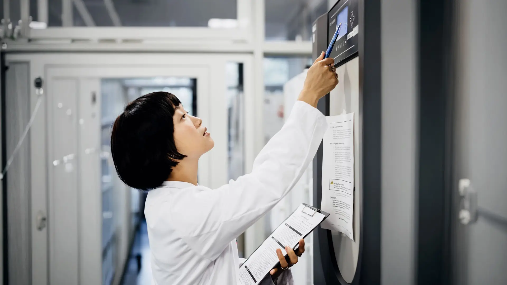 A laboratory technician holding printed documentation while checking a lab instrument, illustrating manual record-keeping